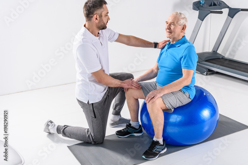 rehabilitation therapist assisting senior man exercising on fitness ball