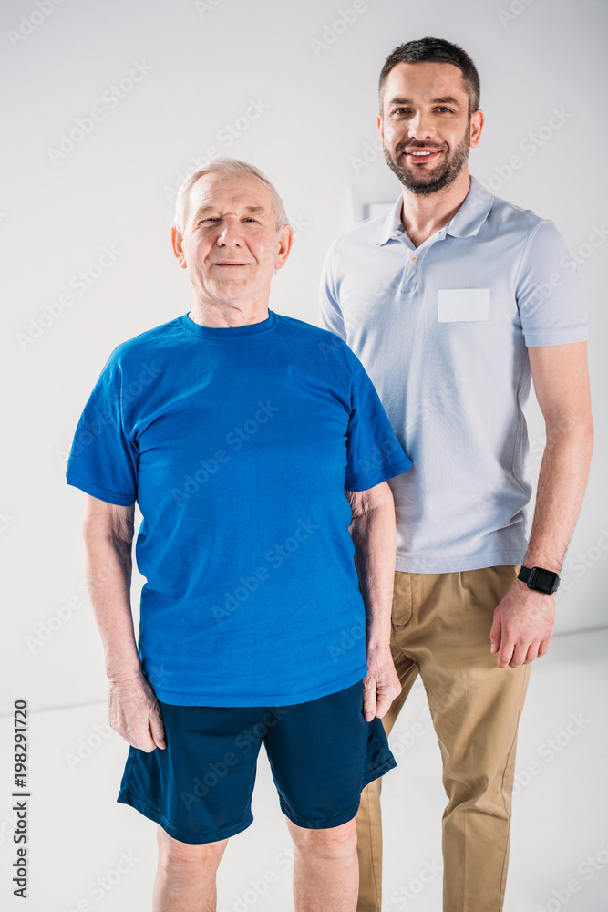 portrait of smiling senior man and rehabilitation therapist looking at camera on grey backdrop portrait of smiling senior man and rehabilitation therapist looking at camera on grey backdrop
