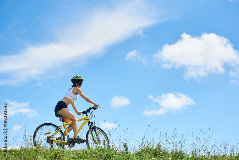 Back view of athlete young biker riding on yellow mountain bike against blue sky with clouds, Woman wearing helmet, enjoying valley view on sunny day. Outdoor sport activity. Copy space