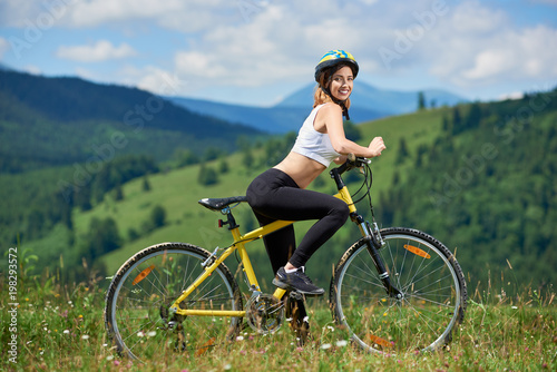 Sporty slim girl biker posing with yellow bicycle in the mountains on sunny day, wearing helmet. Smiling to the camera. Outdoor sport activity, lifestyle concept