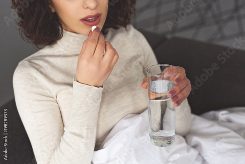 cropped image of sick woman taking pill and holding glass of water at home photo