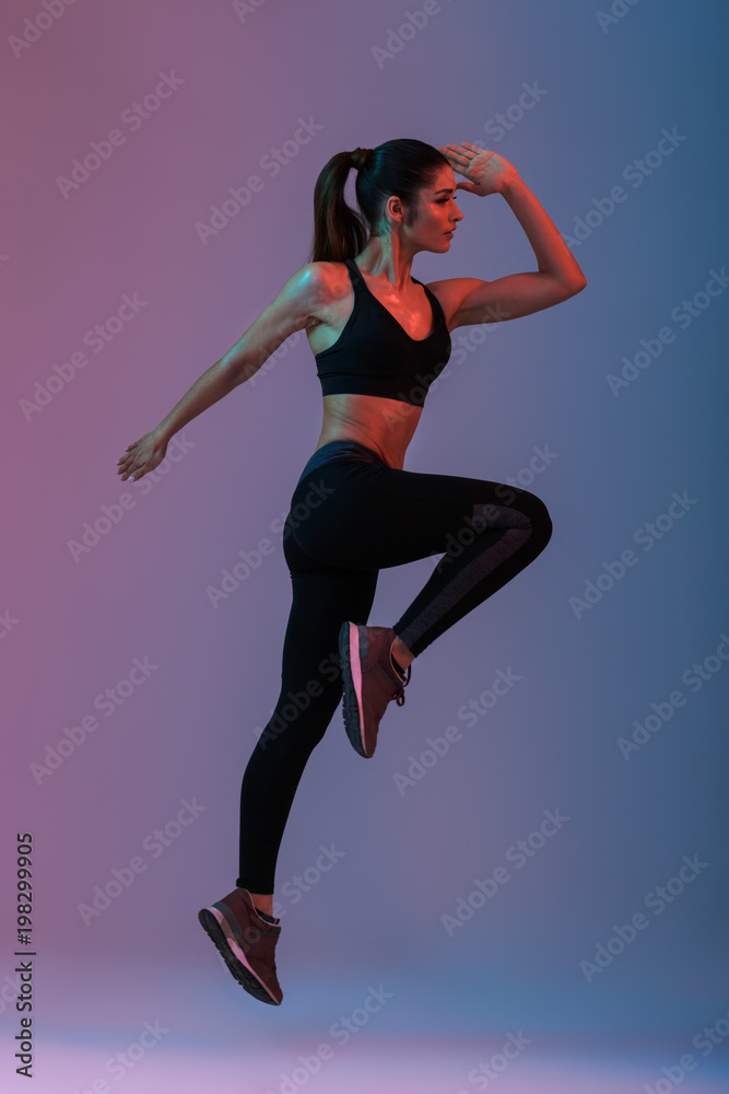 Full length portrait in profile of female fitness instructor wearing black tracksuit doing aerobics in gym, isolated over purple background