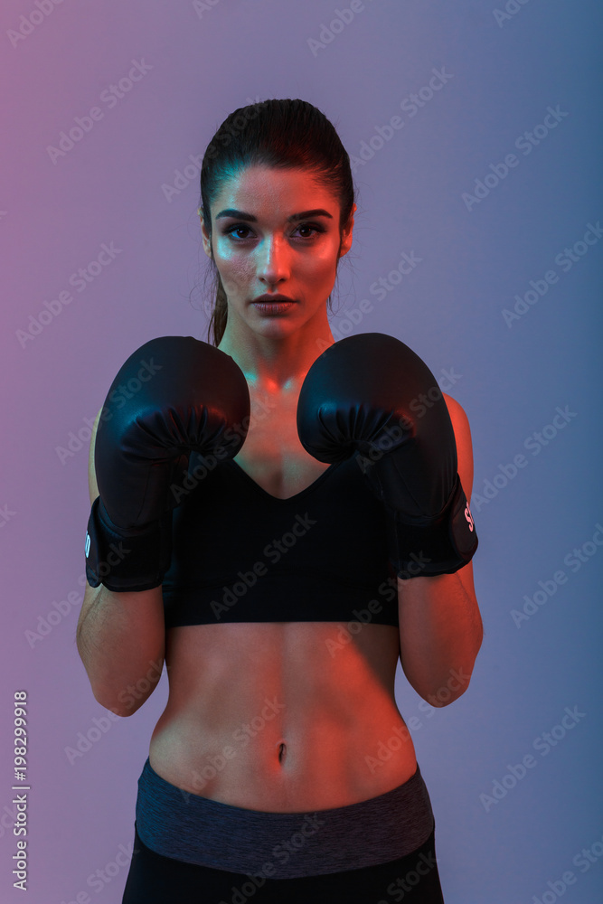 Feminine woman fighter 20s in sportswear and black boxing gloves standing in defense position while training, isolated over purple background