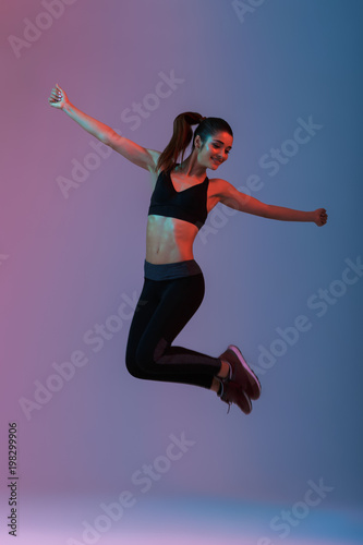 Full length portrait of happy woman wearing black tracksuit jumping with hands aside during aerobics in gym, isolated over purple background