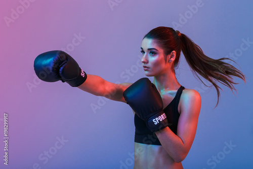 Feminine woman fighter 20s in sportswear and black boxing gloves throwing punches while training, isolated over dark background photo