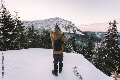 Professional photographer with warm clothes taking photos with her camera from a snow covered trees and moountains at sunrise