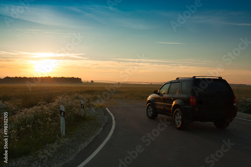 Beautiful Landscape With Black Car On Asphalt Road Under Blue Sky With Sunset Of Sun In Early Dawn Summer.