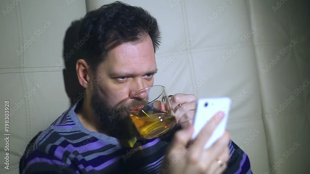 a bearded man watching news on the smartphone's screen and drinking tea and responding to the call. From a surprise spits out tea from his mouth