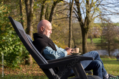 Senior man relaxing in chair outside with mobile phone and cup of coffee