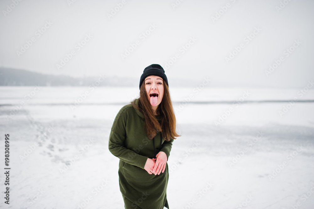 Funny girl wear on long green sweatshirt, jeans and black headwear shows a tongue at frozen lake in winter day. Funny girl wear on long green sweatshirt, jeans and black headwear shows a tongue at frozen lake in winter day.