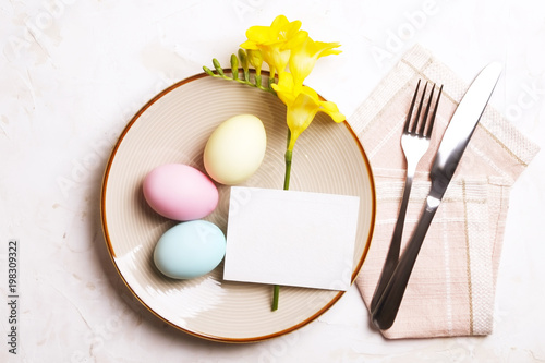 Easter table setting composition: colorful eggs, plate, fork & knife silverware on napkin, yellow freesia flower on white stucco background. celebrating festive Top view, copy space, close up