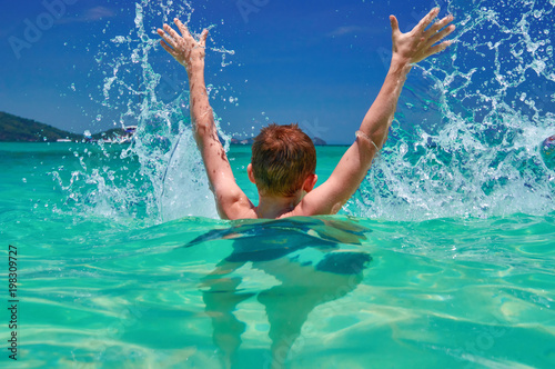 Child splashing in sea. Boy (10 years old) hands up in turquoise water tropical sea. Seascape with bright sky and sea. Back view.