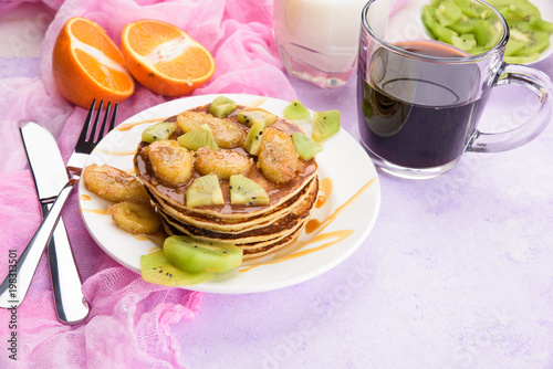 Homemade breakfast on a marble violet table. Stack of pancakes with a grilled bananas and a caramel. Glass of juice, orange, kiwi slices.Healthy breakfast concept, flat lay, top view