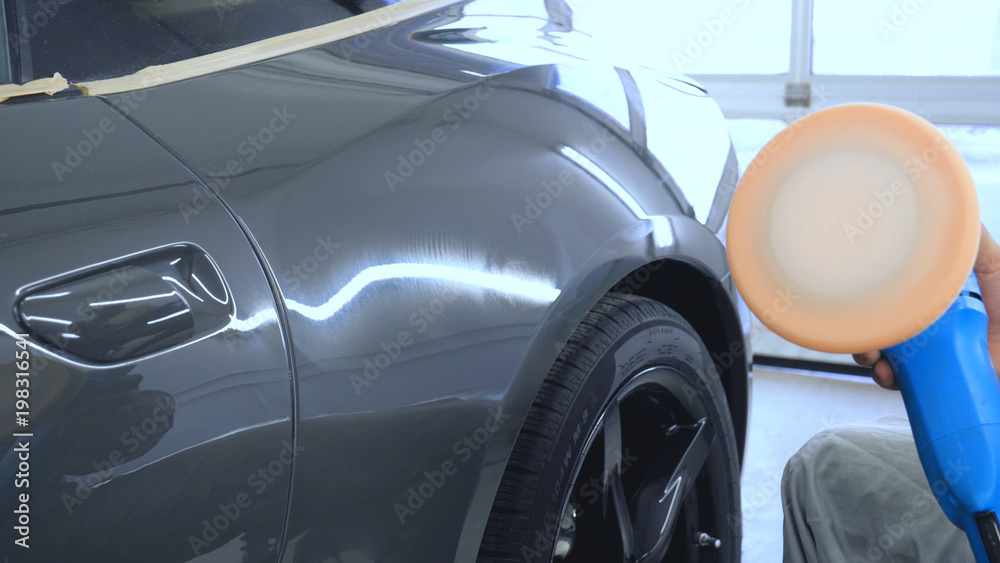 A man in a special suit polishes a gray car body, a tool for polishing cars, into a workshop.