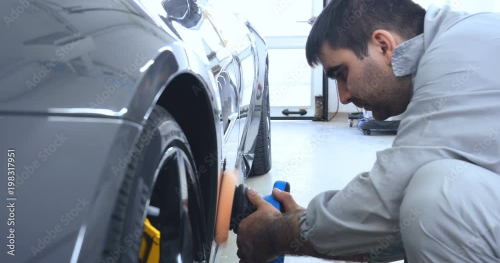 A man in a special suit polishes a gray car body, a tool for polishing cars, into a workshop. A man in a special suit polishes a gray car body, a tool for polishing cars, into a workshop.