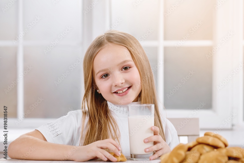 Smiling little girl is drinking milk from transparent glass with delicious cookies,which lay on brown patterned plate. Child wears white sweatshirt and has long blonde hair. Window on background. Smiling little girl is drinking milk from transparent glass with delicious cookies,which lay on brown patterned plate. Child wears white sweatshirt and has long blonde hair. Window on background.