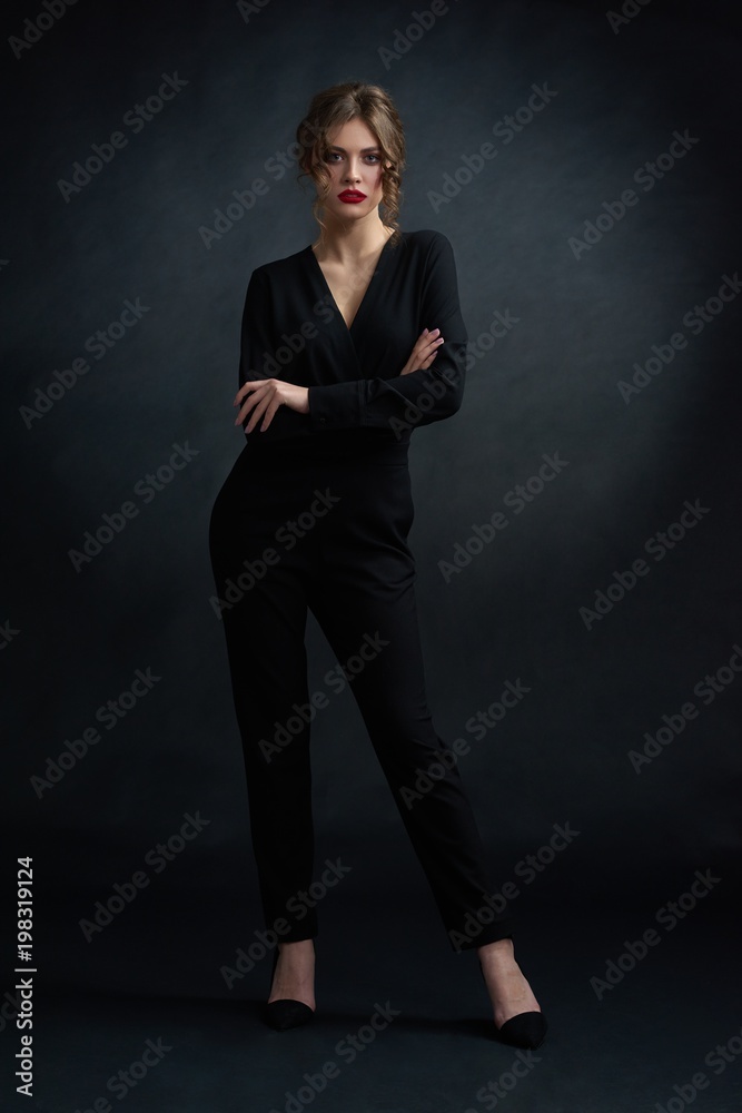 Studio frontview of young confident woman wearing black stylish suit and posing in front of camera. Model wearing bright red lipstick, day make up and hairstyle with curles.