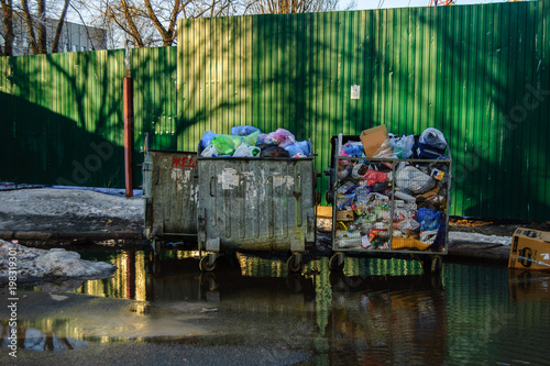 Overflowing garbage dumpsters by green fence in the trash area near the apartment building