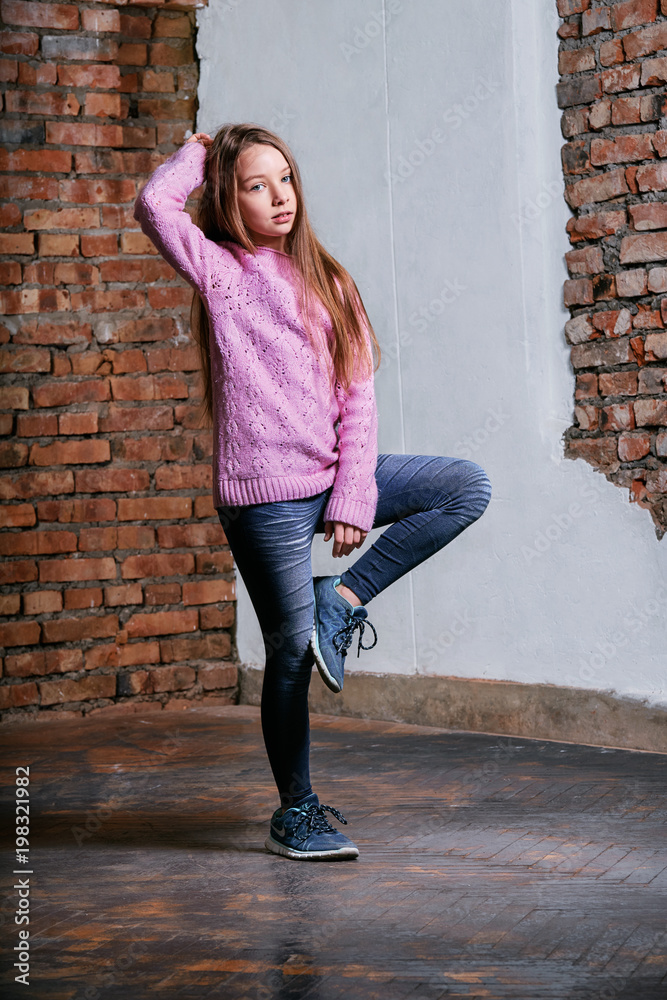 Portrait little fashion kid girl on brick wall background. Model tests young caucasian teen girl in pink sweater posing at studio loft,urban,street style. Teenager raising hand up holding hair.