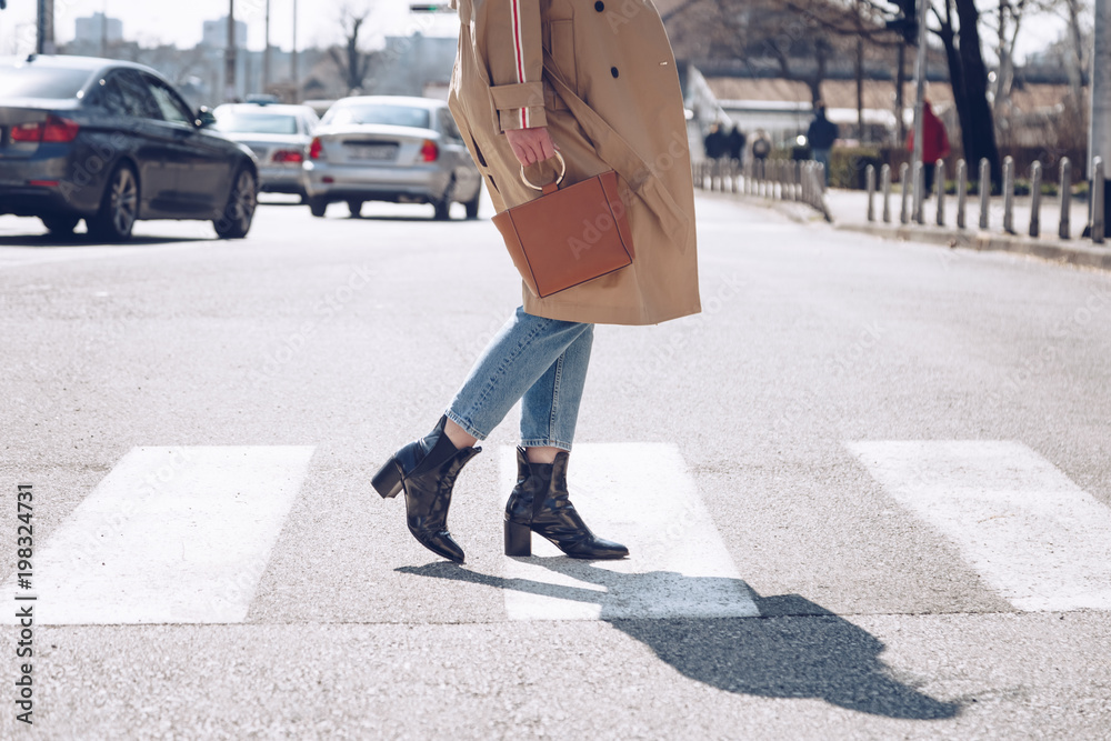 street style portrait of an attractive woman wearing a beige trench coat, denim jeans, ankle boots, cat eye sunglasses and a metallic handle brown tote bag. fashion outfit perfect for sunny spring day