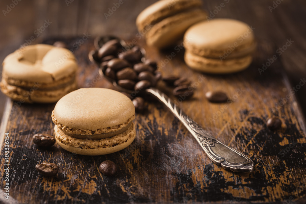 French coffee macaroons with ganache filling and spoon with coffee beans on old wooden board on vintage background. Holidays food concept. French coffee macaroons with ganache filling and spoon with coffee beans on old wooden board on vintage background. Holidays food concept.