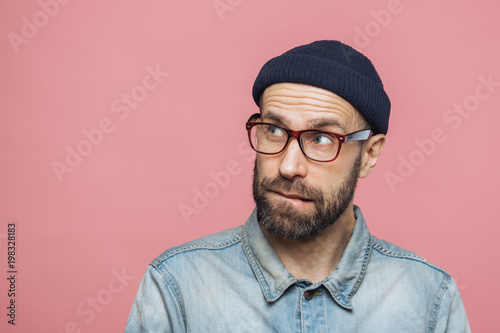 Portrait of unshaven thoughtful middle aged male with pensive expression looks upwards, wears stylish hat, eyewear and denim jacket, isolated over pink background with copy space for your text
