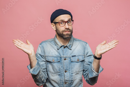 Photography Indoor shot of hesitant bearded man shrugs shoulders, looks uncertain, doubts when makes decision, isolated over pink background