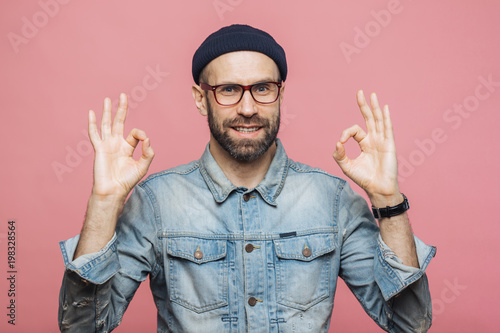 Photography Cheerful bearded male in fashionable clothing, makes ok sign, being satisfied with something, isolated over pink studio background