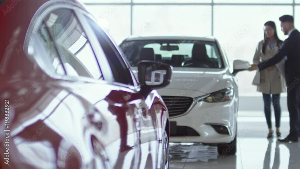 PAN of professional salesman in suit showing new car to young Asian woman at dealership PAN of professional salesman in suit showing new car to young Asian woman at dealership