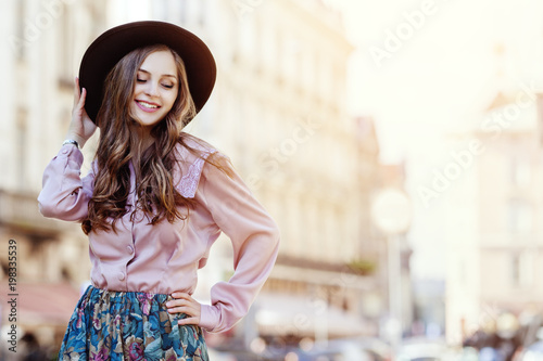 Outdoor portrait of a young beautiful fashionable happywoman posing on a street of the old european city. Model wearing stylish hat, pink blouse, printed skirt. Female fashion concept. Copy space
 photo