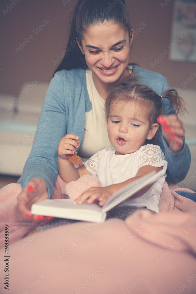 Cheerful mother and her little daughter at home sitting on floor and reading book together. Cheerful mother and her little daughter at home sitting on floor and reading book together.