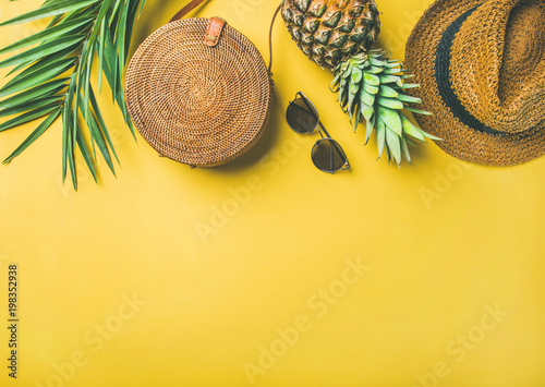 Colorful summer female fashion outfit flat-lay. Straw hat, bamboo bag, sunglasses, palm branches, fresh pineapple over yellow background, top view, copy space. Summer fashion, holiday travel concept
