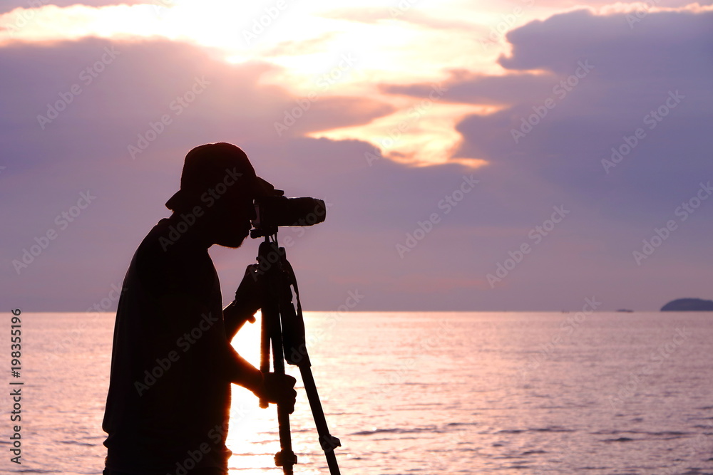 Silhouette photographer taking photo of sunset light with tripod at sea in Thailand. The sky have purple and pink color tone.