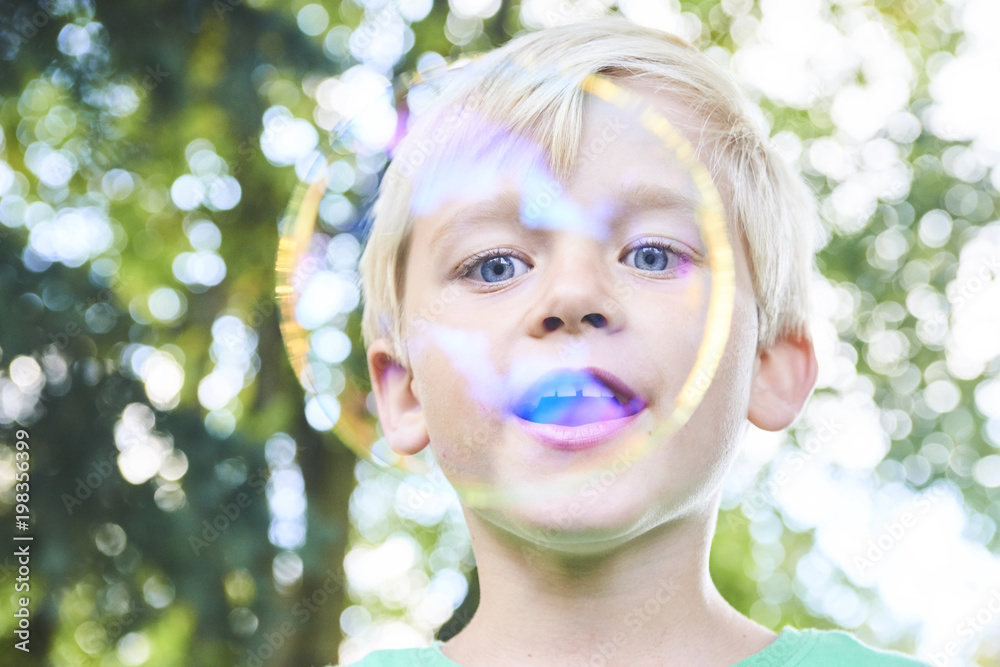 Young boy blowing bubbles with greenery background. Selective focus.
Young boy blowing bubbles with greenery background. Selective focus.
