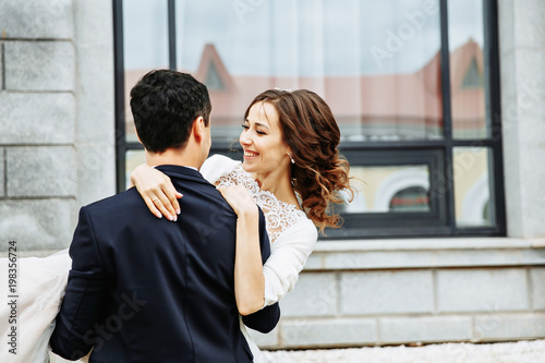 the groom holds his bride in his arms and they dance near the building with beautiful windows