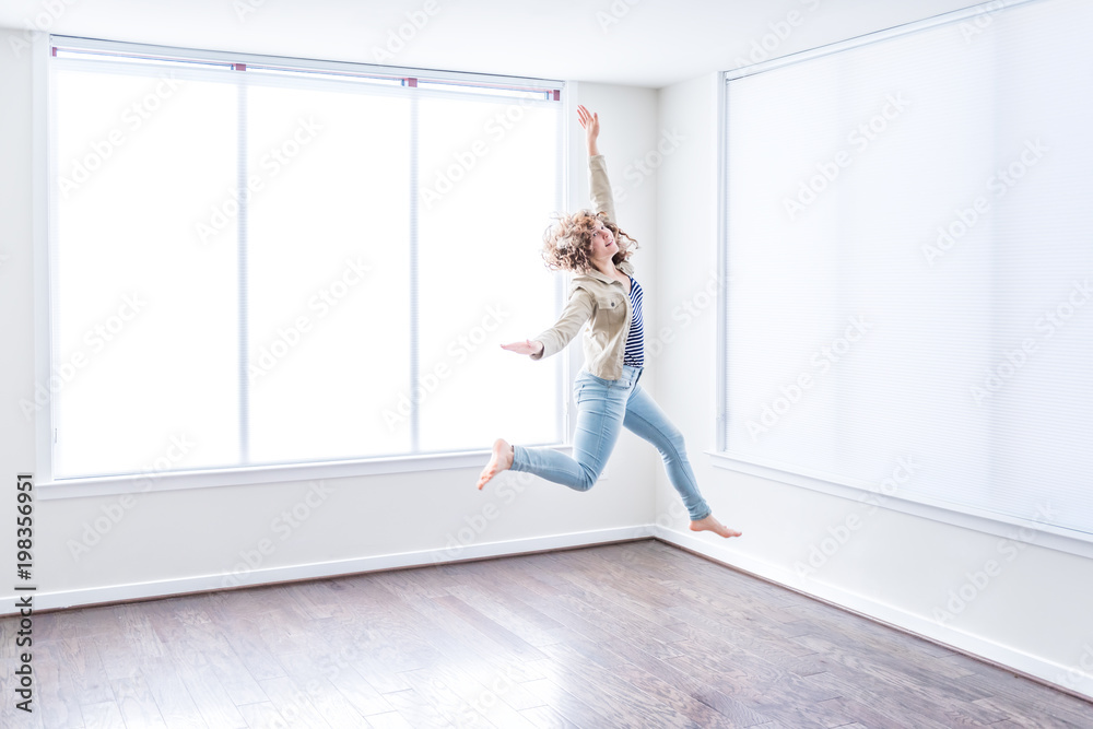 One young happy woman jumping up in empty modern new room with hardwood floors and large sunny windows in apartment