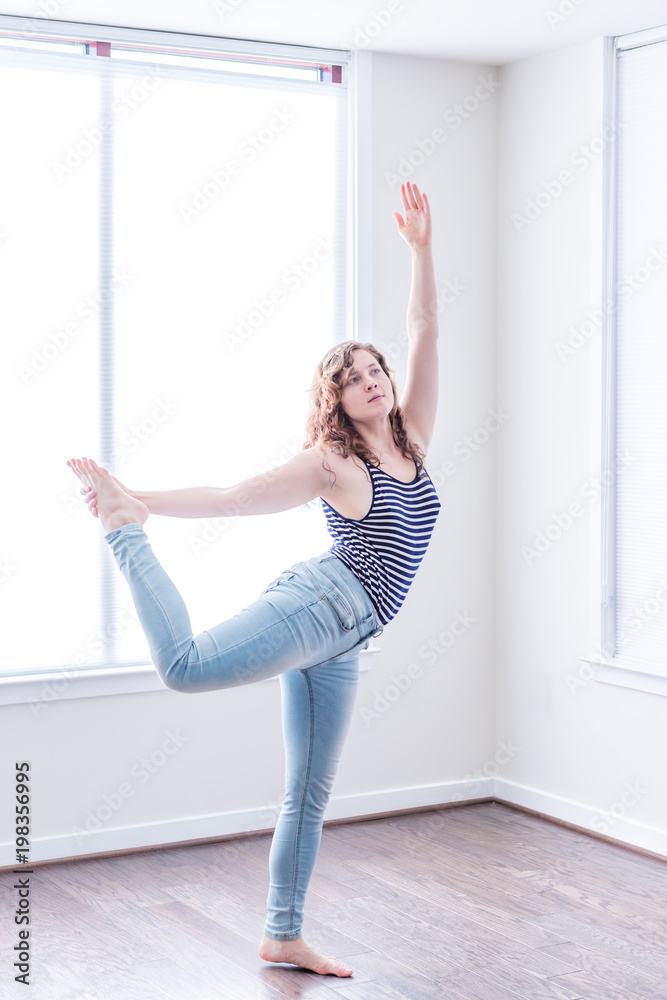 Young happy woman stretching dancing in empty modern new room with hardwood floors and large sunny windows in apartment
