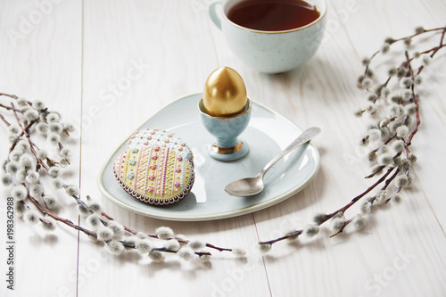Close-up photo of Easter golden egg on wooden background with willows branches and cup of tea on plate with cookies