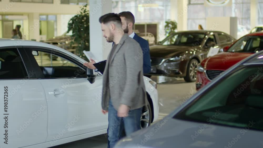 PAN of auto salesman in suit opening door of demo car and talking with male client choosing car at dealership PAN of auto salesman in suit opening door of demo car and talking with male client choosing car at dealership