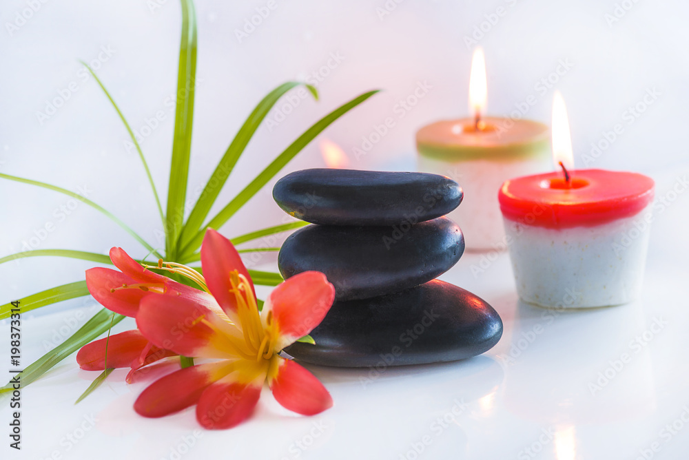 Beautiful spa compostion with black massage stones, red flowers and burning candles on white glossy background