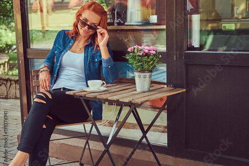 A beauty redhead girl drinks coffee, sitting near the coffee shop.