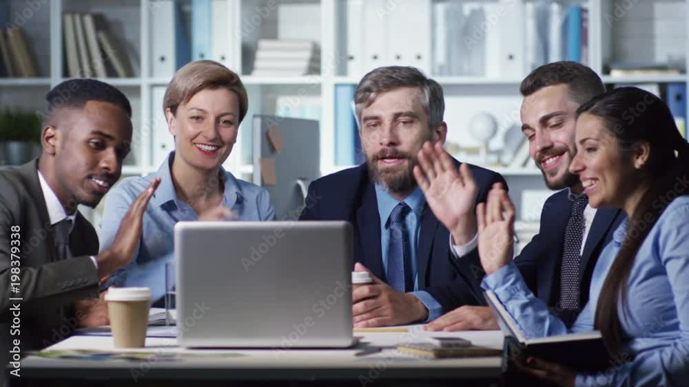 Group of colleagues sitting together at desk and waving hands to their partner during online meeting Group of colleagues sitting together at desk and waving hands to their partner during online meeting