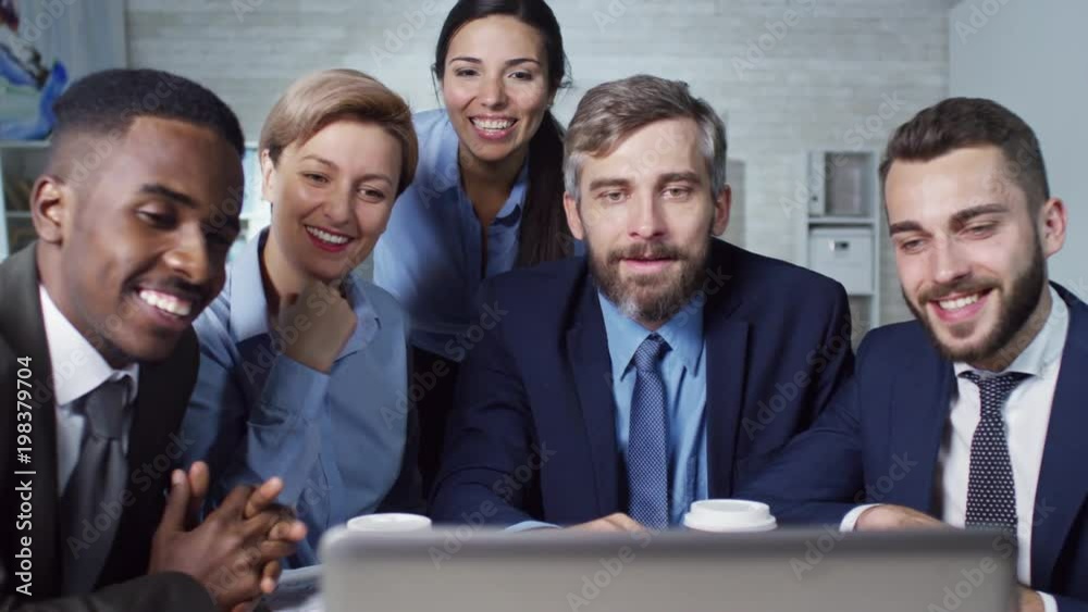 Group of colleagues sitting together in front of laptop computer, smiling and waving hands to their partner during online meeting Group of colleagues sitting together in front of laptop computer, smiling and waving hands to their partner during online meeting
