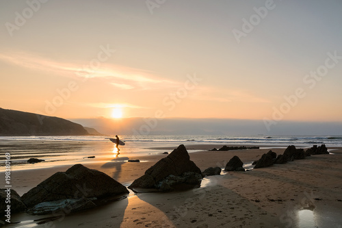 Surfer walking backlit on the beach at magical golden sunset with sharp rocks
