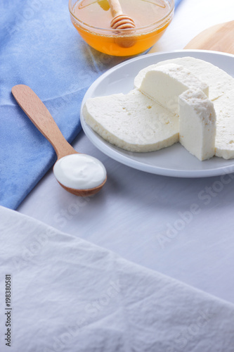 Cheese, honey and sour cream, feta in a white plate, soft cheese on a white background, wooden spoon with sour cream, French breakfast, blue napkin, honey in glassware, art