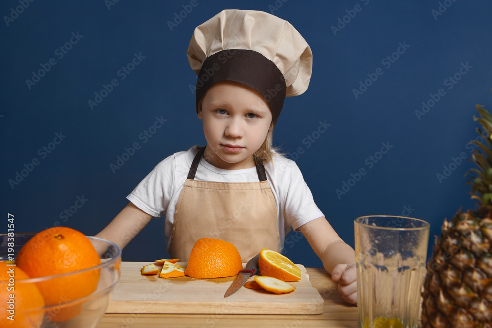 Isolated shot of serious male child chef with blue eyes and blonde hair making fruit fresh or salad, posing at blank wall with bowl of oranges, glass, pineapple and cooking board with knife on table Isolated shot of serious male child chef with blue eyes and blonde hair making fruit fresh or salad, posing at blank wall with bowl of oranges, glass, pineapple and cooking board with knife on table