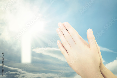 Concept of Christian prayer - folded hands for prayer against a blue sky and a shining cross sign (mixed)