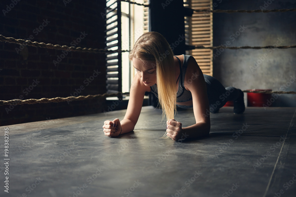 Beautiful young blonde woman in sports clothes exercising indoors, doing elbow plank inside boxing ring, testing endurance, having concentrated focused look. Athletic sporty girl training in gym