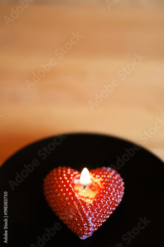 Close up of red heart shape burning candle on black candle holder at shallow depth of field on brown orange blur background with copy space for text. 