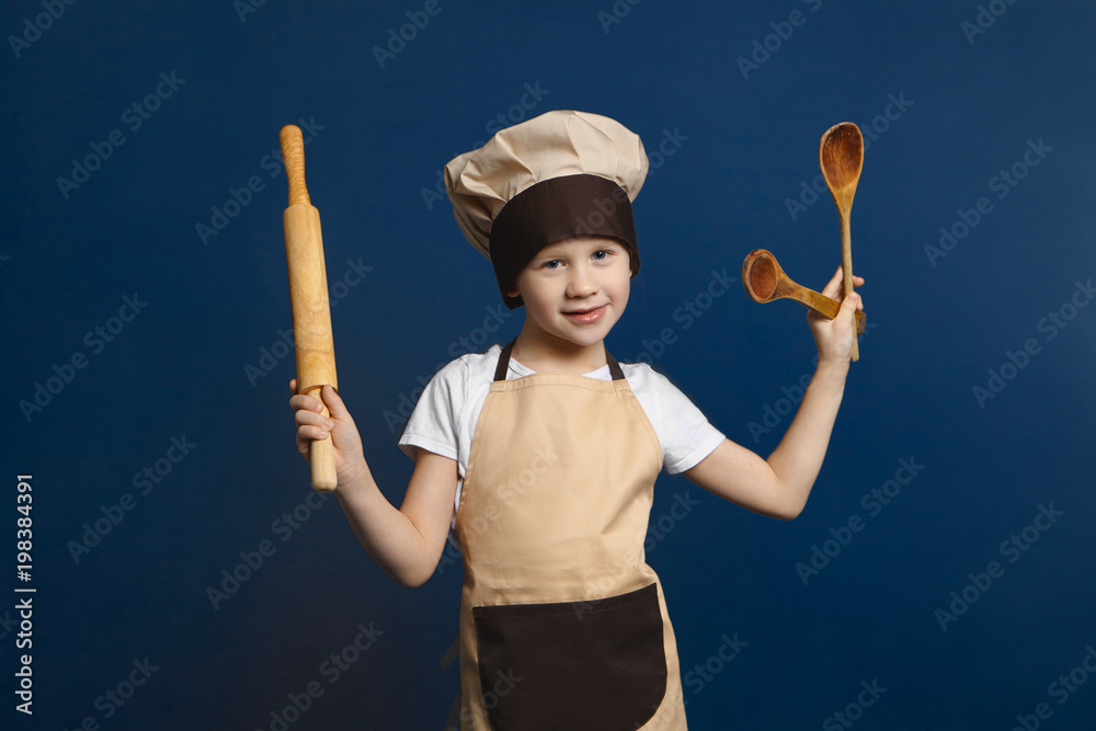 Fototapeta premium Children, cooking, cuisine and food concept. Picture of funny little boy wearing chef uniform standing at blank wall, holding rolling pin in one hand and two wooden spoons in other, smiling at camera